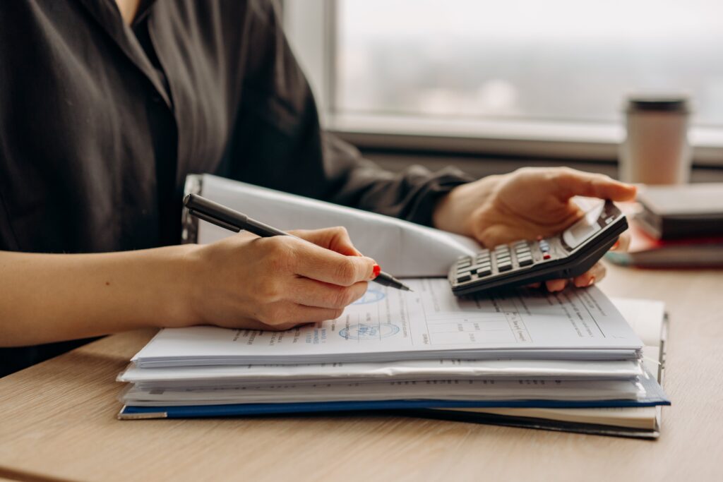 educação financeira pessoa mostrando os braços e mãos e em uma das mãos uma caneta e na outra uma calculadora sobre um caderno de anotações.